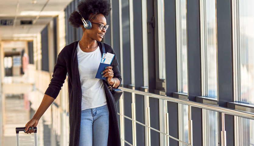 Young african american female passanger in casual clothes and headphones is in airport with baggage. holding passport and boarding card